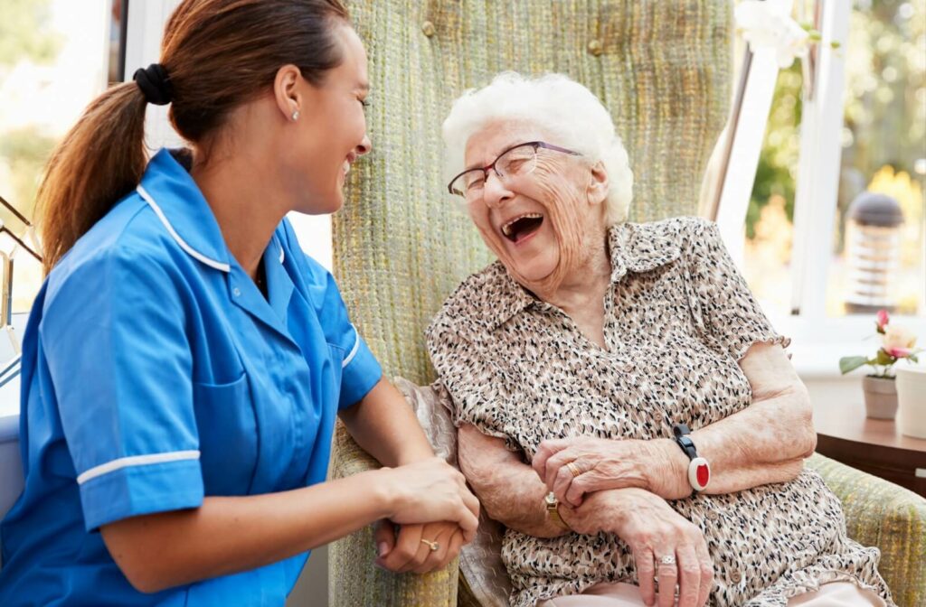 An senior adult woman in a memory care facility sitting on a chair smiling and having a conversation with a nurse.