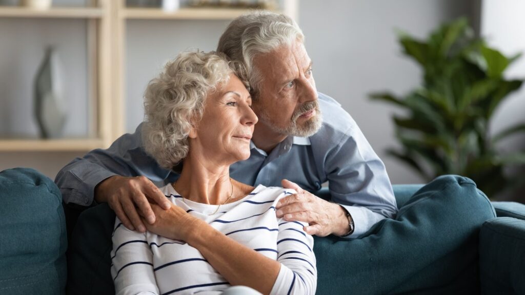 An older couple with grey hair is sitting on the couch together, staring out the window in peaceful contemplation.