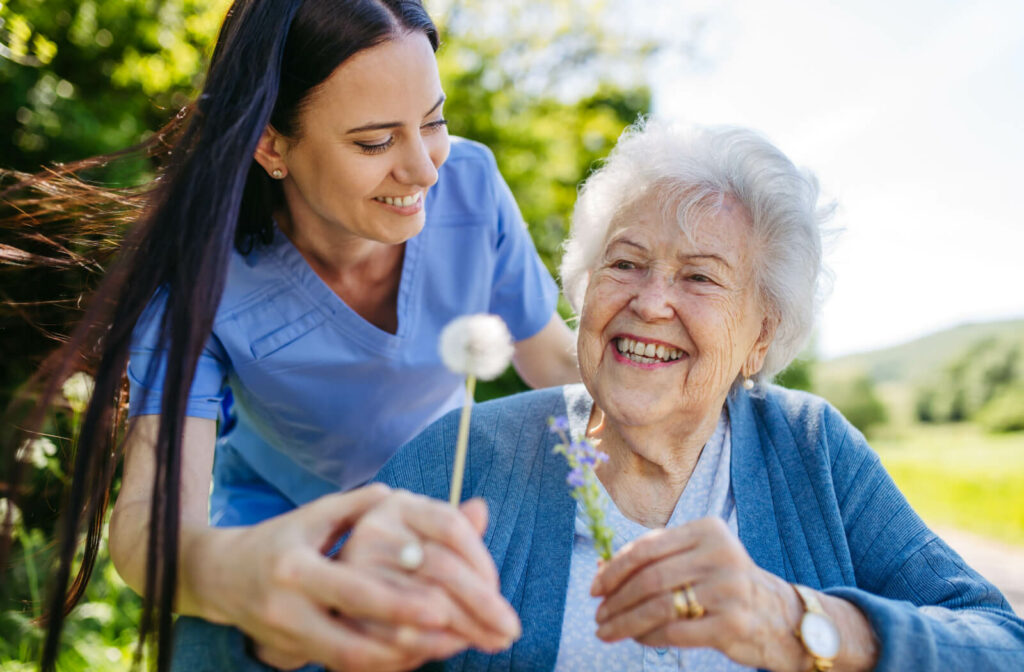 A caregiver helping an older adult in memory care pick a dandelion while on an outdoor walk together.